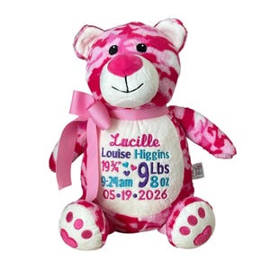 Puede incluir: Un oso de peluche con estampado de camuflaje rosa y blanco, con una cinta de satén rosa alrededor del cuello. El vientre blanco está bordado con el nombre Lucille Louise Higgins y detalles de nacimiento: 19 3/4 pulgadas, 9 lbs 8 oz, 9:24am y 05-19-2026.