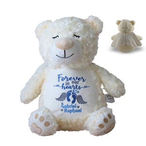 May include: Cream-colored plush teddy bear with embroidered text "Forever in our hearts" and the names Gabriel and Raphael. The bear has closed eyes and a small angel wing design on the back. A second bear with angel wings is also shown.