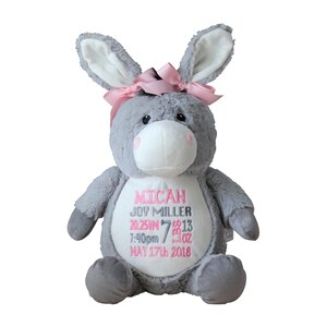 May include: A gray plush donkey stuffed animal with a pink bow on its head. The donkey has a white belly with embroidered text in pink: "MICAH JOY MILLER 20.25IN 7:40pm 7 OZ MAY 17th 2016".