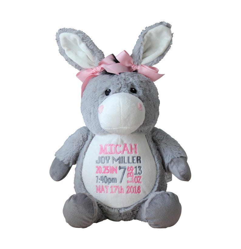 May include: A gray plush donkey stuffed animal with a pink bow on its head. The donkey has a white belly with embroidered text in pink: "MICAH JOY MILLER 20.25IN 7:40pm 7 OZ MAY 17th 2016".