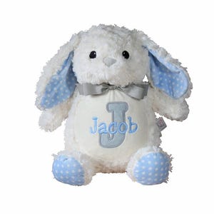 May include: A white plush bunny with blue polka dot accents on the ears and feet. The bunny has a gray bow and a cream-colored belly with the name "Jacob" and the letter "J" embroidered in blue.