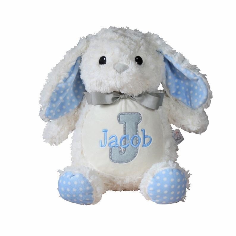 May include: A white plush bunny with blue polka dot accents on the ears and feet. The bunny has a gray bow and a cream-colored belly with the name "Jacob" and the letter "J" embroidered in blue.