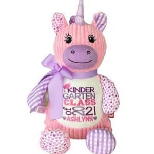 May include: A pink plush unicorn with a purple horn, ears, and accents. The unicorn has a ribbon around its neck and a white shirt with the text "KINDERGARTEN CLASS 2021 ASHLYNN". The unicorn's feet and arms have patterned designs.
