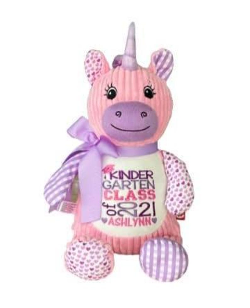May include: A pink plush unicorn with a purple horn, ears, and accents. The unicorn has a ribbon around its neck and a white shirt with the text "KINDERGARTEN CLASS 2021 ASHLYNN". The unicorn's feet and arms have patterned designs.