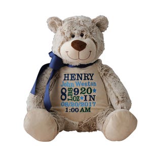 May include: A plush teddy bear with a brown nose and a blue ribbon around its neck. The bear has embroidered text on its belly, including a name, weight of 8 lbs 9 oz, and date and time of birth. The bear is light brown.