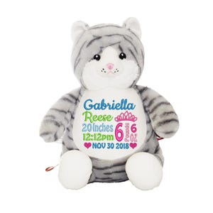 May include: A gray and white plush cat with embroidered details. The cat has the name "Gabriella Reese", measurements of 20 inches, 6 pounds, and the date Nov 30 2018. The cat has white paws and a pink nose.