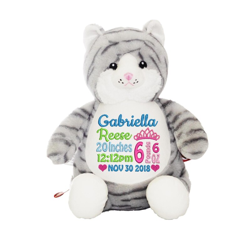 May include: A gray and white plush cat with embroidered details. The cat has the name "Gabriella Reese", measurements of 20 inches, 6 pounds, and the date Nov 30 2018. The cat has white paws and a pink nose.