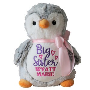 May include: A plush penguin toy with gray and white fur, an orange beak and feet, and black eyes. It has a pink ribbon around its neck and a white belly embroidered with "Big Sister" and a name in purple and pink.