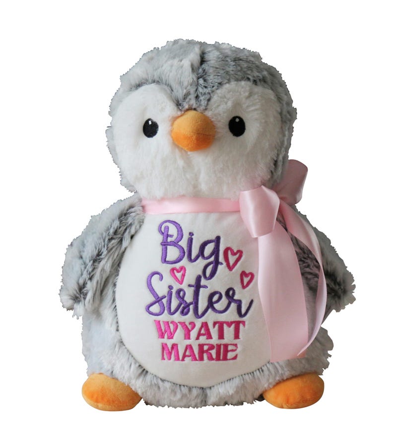 May include: A plush penguin toy with gray and white fur, an orange beak and feet, and black eyes. It has a pink ribbon around its neck and a white belly embroidered with "Big Sister" and a name in purple and pink.