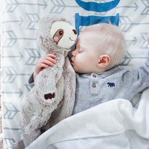 Pu&ograve; includere: Un bambino che dorme in una culla, che abbraccia un peluche di bradipo. Il bambino indossa una maglietta grigia a maniche lunghe con un'emblema di orso. La culla ha un lenzuolo fantasia con frecce e una coperta bianca.