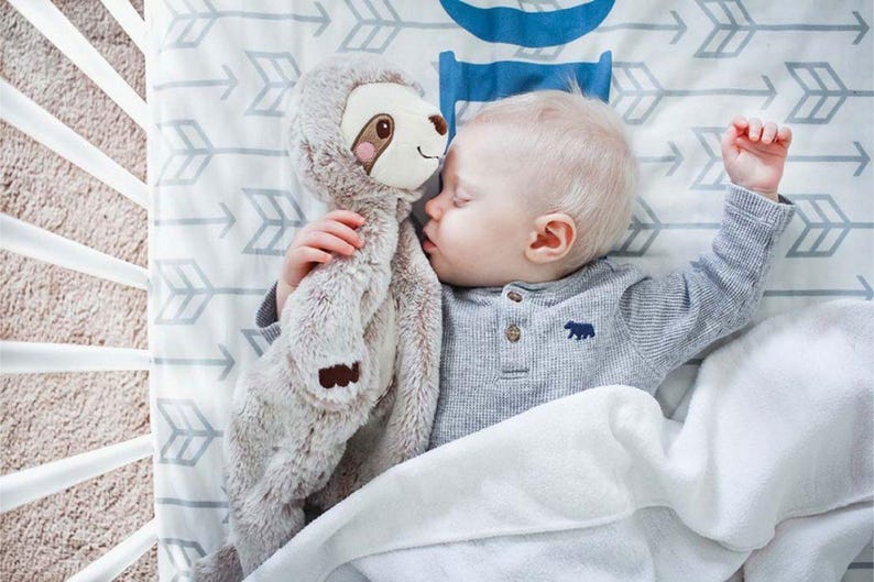 Pu&ograve; includere: Un bambino che dorme in una culla, che abbraccia un peluche di bradipo. Il bambino indossa una maglietta grigia a maniche lunghe con un'emblema di orso. La culla ha un lenzuolo fantasia con frecce e una coperta bianca.