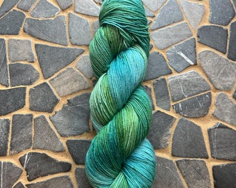 May include: A skein of hand-dyed yarn in shades of green and teal. The yarn is twisted into a compact, elongated shape, showcasing the color variations. The yarn is displayed against a background of irregularly shaped gray stones.