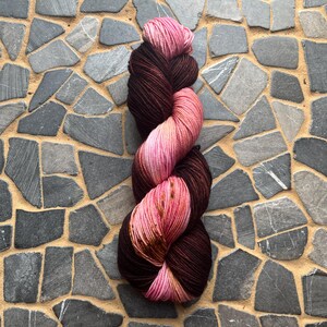 May include: A skein of hand-dyed yarn in shades of pink and brown. The yarn is twisted into a compact bundle, with the colors transitioning from a deep brown to a light pink. The yarn is likely made of wool or a similar fiber, perfect for knitting or crocheting.
