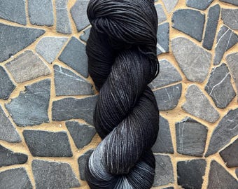 May include: A skein of black yarn with subtle gray and white gradients. The yarn is tightly wound, showcasing a smooth texture. The background is a mosaic of gray and tan stones, providing a neutral backdrop for the yarn.