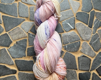 May include: A skein of hand-dyed yarn in a blend of pink, white, and orange hues. The yarn has a speckled appearance with hints of purple and yellow. The yarn is tightly wound and ready for knitting or crocheting projects.