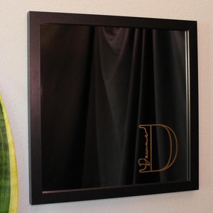 Może przedstawiać: Czarne lustro w ramie z pozłacaną literą D i napisem "Dawson" wewnątrz litery. Lustro wisi na białej ścianie.