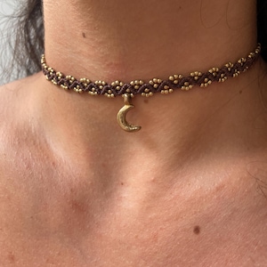 Può includere: Una collana girocollo in macramè marrone con perline color oro e un ciondolo a forma di mezzaluna. La collana è indossata intorno al collo.