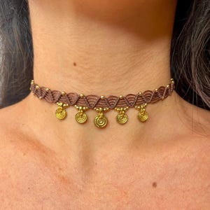Op de afbeelding: Een bruine macramé choker ketting met goudkleurige muntbedels. De ketting wordt om de nek gedragen en heeft een geweven ontwerp met kleine gouden kralen. De gouden muntbedels hangen onder de geweven band.