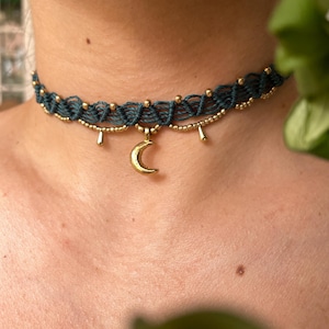 Può includere: Una collana girocollo in macramè verde scuro con perline color oro e un ciondolo a forma di mezzaluna. La collana è finemente intrecciata e presenta piccoli accenti dorati pendenti, creando un'estetica delicata e bohémien.