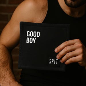 K&ouml;nnte beinhalten: Schwarzes quadratisches Textil mit den Worten "GOOD BOY" in wei&szlig;en Blockbuchstaben. Ein kleineres wei&szlig;es Etikett lautet "SPIT". Das Textil wird vor einer Person gehalten.