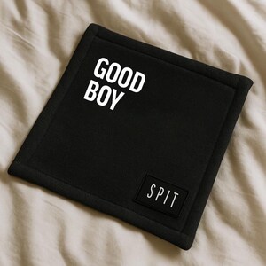 K&ouml;nnte beinhalten: Schwarzes quadratisches Hundebett mit den Worten "GOOD BOY" in gro&szlig;en wei&szlig;en Buchstaben. Ein kleines rechteckiges Patch mit dem Wort "SPIT" ist in der unteren rechten Ecke angen&auml;ht. Das Bett liegt auf einer beigen Oberfl&auml;che.