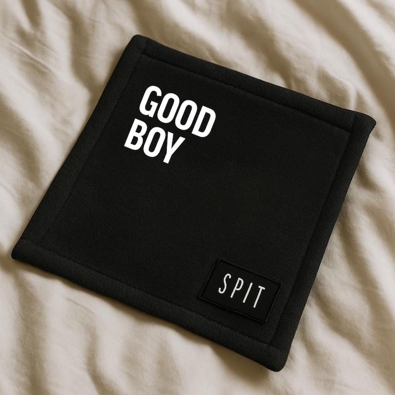 K&ouml;nnte beinhalten: Schwarzes quadratisches Hundebett mit den Worten "GOOD BOY" in gro&szlig;en wei&szlig;en Buchstaben. Ein kleines rechteckiges Patch mit dem Wort "SPIT" ist in der unteren rechten Ecke angen&auml;ht. Das Bett liegt auf einer beigen Oberfl&auml;che.