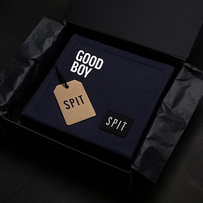 K&ouml;nnte beinhalten: Ein marineblaues quadratisches Textil mit den wei&szlig;en W&ouml;rtern "GOOD BOY" und "SPIT". Ein braunes Etikett mit "SPIT" ist angebracht. Das Textil befindet sich in einer schwarzen Box.