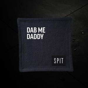 May include: A square, dark blue fabric coaster with the text "DAB ME DADDY" in white block letters. A smaller black rectangle with the word "SPIT" is in the lower right corner.