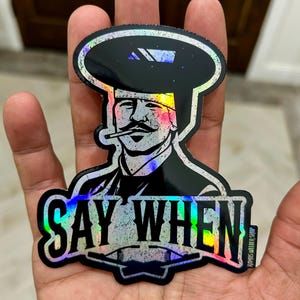 May include: A holographic sticker featuring a stylized illustration of a man with a mustache and a hat, smoking a cigarette. The sticker has the text "SAY WHEN" in bold letters. The sticker is held in a hand.