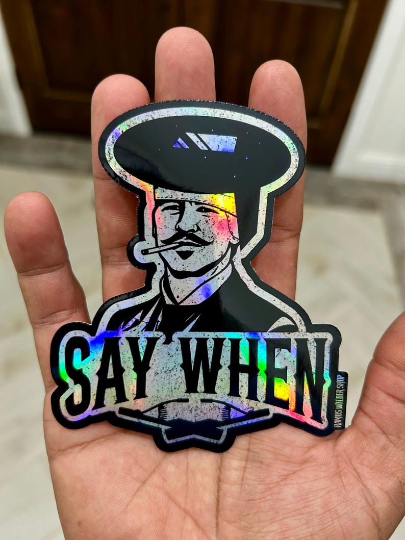 May include: A holographic sticker featuring a stylized illustration of a man with a mustache and a hat, smoking a cigarette. The sticker has the text "SAY WHEN" in bold letters. The sticker is held in a hand.