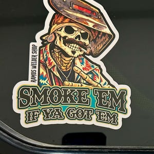 Op de afbeelding: Een sticker met een skeletlasser met een snor, die een lashelm en een kleurrijk shirt draagt. De sticker heeft de tekst "SMOKE EM IF YA GOT EM" en "RAMOS WELDER SHOP".