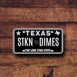 May include: Black and white sticker with a Texas license plate design. It says "Texas" with a star and "STKN Dimes" with a Texas outline. The bottom says "The Lone Star State."