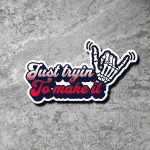 May include: A sticker with a white skeleton hand making a shaka sign. The text "Just tryin' To make it" is in red and blue letters with a white outline.