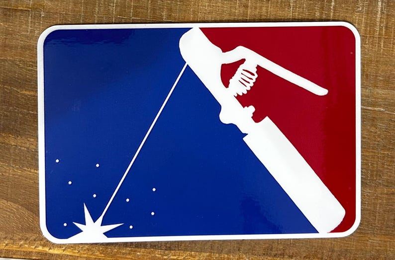 K&ouml;nnte beinhalten: Eine wei&szlig;e Silhouette eines Schwei&szlig;brenners mit Funken, die aus dem Ende fliegen, vor einem blauen und roten Hintergrund. Das Design &auml;hnelt einem Baseball-Teamlogo.
