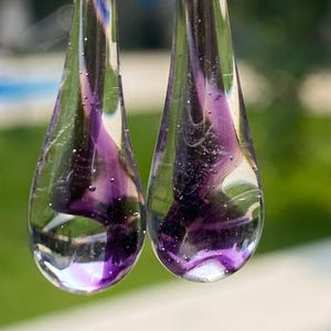 May include: A pair of clear glass teardrop-shaped earrings featuring internal swirls of deep purple and violet pigment. The transparent glass contains small air bubbles suspended within the material, creating a layered effect inside each pendant.
