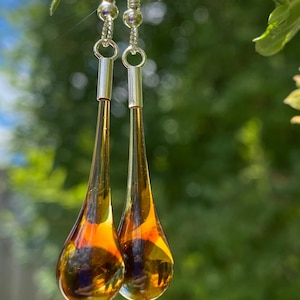 May include: A pair of dangling earrings featuring long, teardrop-shaped amber-colored glass pendants. The pendants are suspended from silver-toned metal hooks and cylindrical connectors, showcasing a smooth, translucent finish with warm golden hues.
