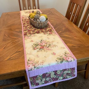 May include: A rectangular table runner featuring a patchwork design with cream fabric printed with pink and red floral patterns, accented by sections of dark maroon floral fabric and bordered by a light pink textured fabric trim.