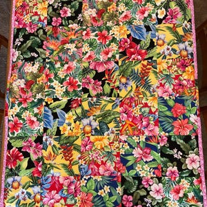 May include: A colorful patchwork quilt featuring a vibrant tropical floral pattern. The quilt showcases squares of fabric with hibiscus, orchids, and other flowers in shades of red, pink, yellow, and blue, bordered by a pink trim.