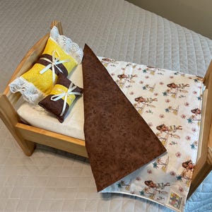 May include: A wooden doll bed with a brown and white patterned blanket and two pillows. One pillow is yellow with white lace and a white ribbon. The other pillow is brown with a yellow ribbon.