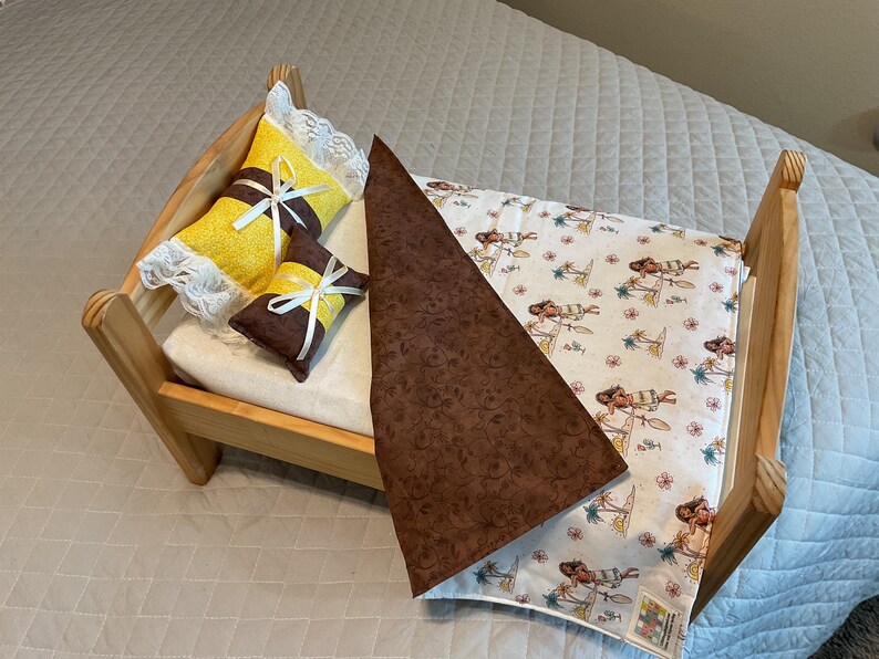 May include: A wooden doll bed with a brown and white patterned blanket and two pillows. One pillow is yellow with white lace and a white ribbon. The other pillow is brown with a yellow ribbon.