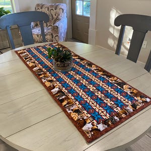 May include: A brown and blue table runner with a geometric pattern and horse silhouettes. The runner is on a round wooden table with a light wood finish.