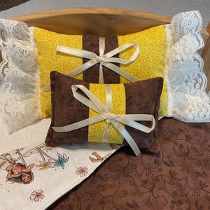 May include: A doll bed with a brown and yellow patterned quilt and two pillows. One pillow is yellow with a brown bow and the other is brown with a yellow bow.