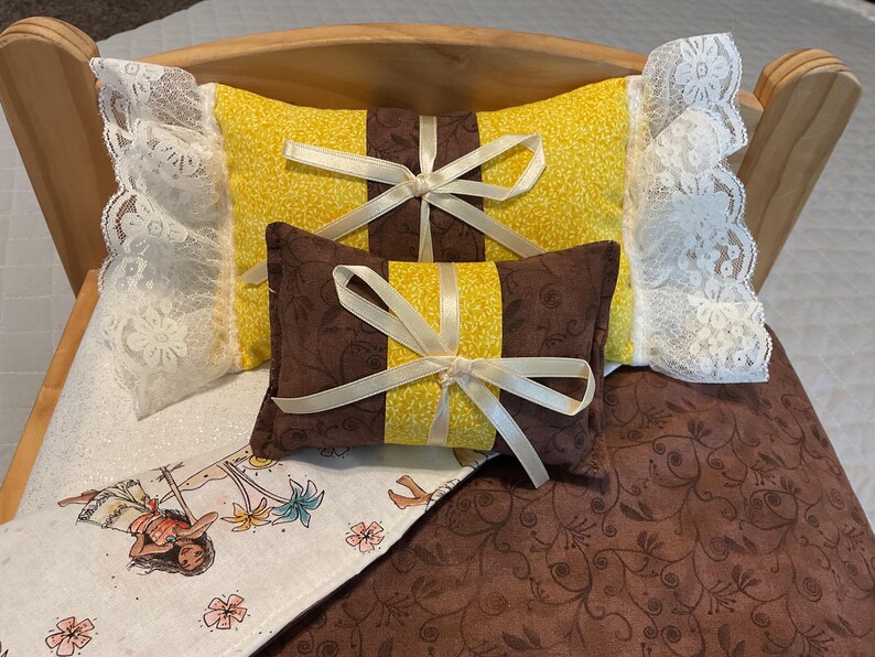 May include: A doll bed with a brown and yellow patterned quilt and two pillows. One pillow is yellow with a brown bow and the other is brown with a yellow bow.