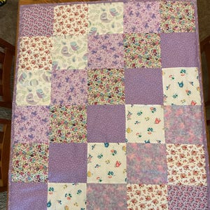 May include: A patchwork quilt with a variety of floral, bird, and polka dot patterns in shades of purple, pink, and white. The quilt is made of square fabric pieces sewn together, with a lavender border. The quilt is likely for a bed or a decorative throw.