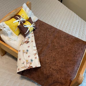 May include: A wooden doll bed with a brown floral patterned comforter and a white comforter with a tropical print. The bed has two yellow pillows with white lace trim and a brown pillow with a white bow.