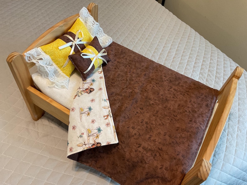 May include: A wooden doll bed with a brown floral patterned comforter and a white comforter with a tropical print. The bed has two yellow pillows with white lace trim and a brown pillow with a white bow.