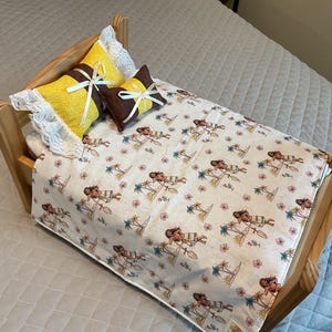 May include: A wooden doll bed with a white and brown patterned bedspread featuring a cartoon character. The bed has two pillows, one yellow with white lace trim and one brown with a white bow.