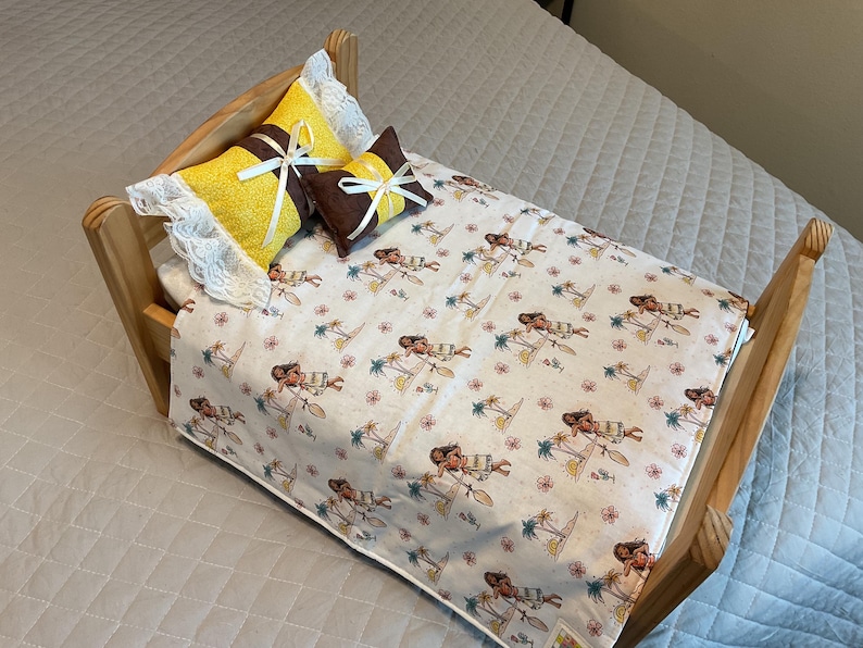 May include: A wooden doll bed with a white and brown patterned bedspread featuring a cartoon character. The bed has two pillows, one yellow with white lace trim and one brown with a white bow.
