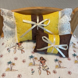 May include: A wooden doll bed with a white lace trim and two pillows. The top pillow is yellow with a brown stripe and a white satin ribbon. The bottom pillow is brown with a yellow stripe and a white satin ribbon. The bed has a white and brown floral patterned sheet.