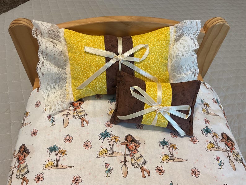 May include: A wooden doll bed with a white lace trim and two pillows. The top pillow is yellow with a brown stripe and a white satin ribbon. The bottom pillow is brown with a yellow stripe and a white satin ribbon. The bed has a white and brown floral patterned sheet.